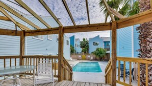 Una terraza de madera con una mesa y sillas de vidrio, una piscina y vistas a casas azules.