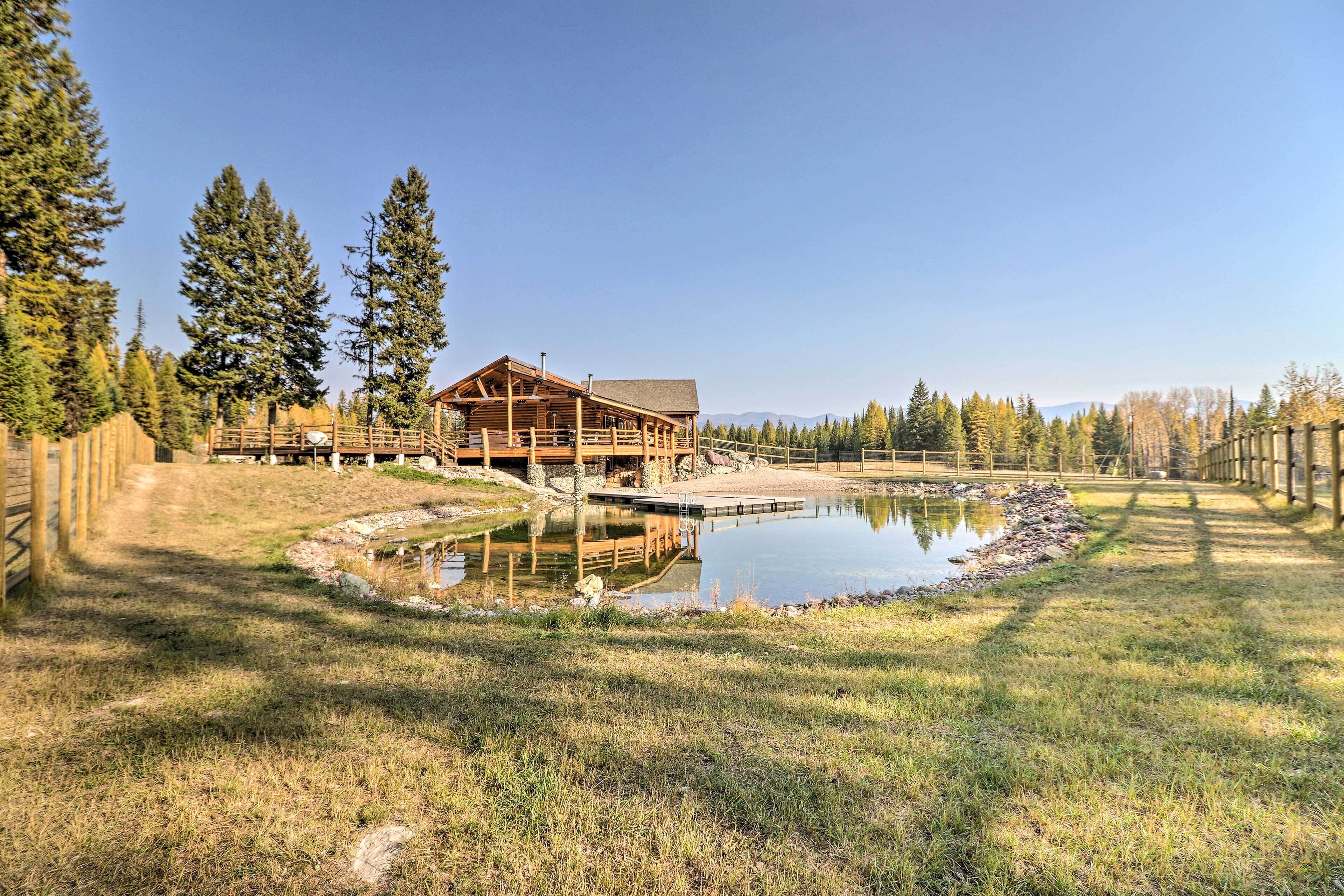 Eine Holzhütte mit Teich, umgeben von hohen Bäumen und blauem Himmel.