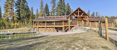 A wooden cabin with a covered deck, surrounded by a fenced area and a pond.