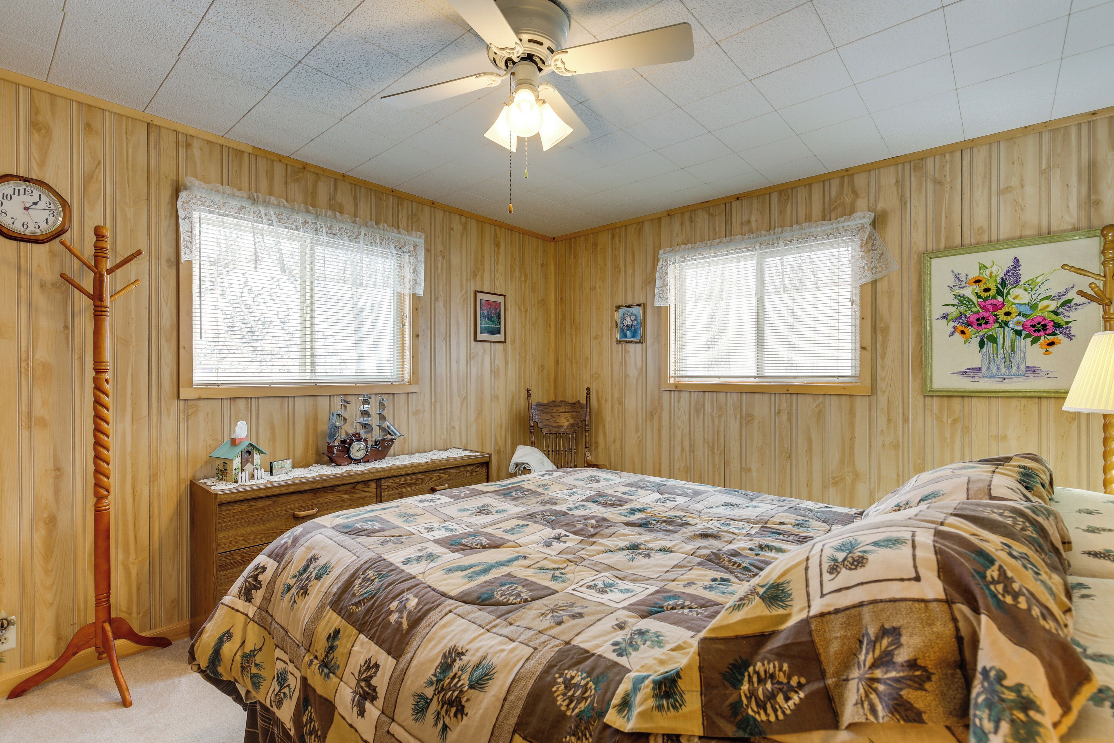 A bedroom with wooden paneling, a bed with a floral quilt, a desk with a clock, and a ceiling fan.