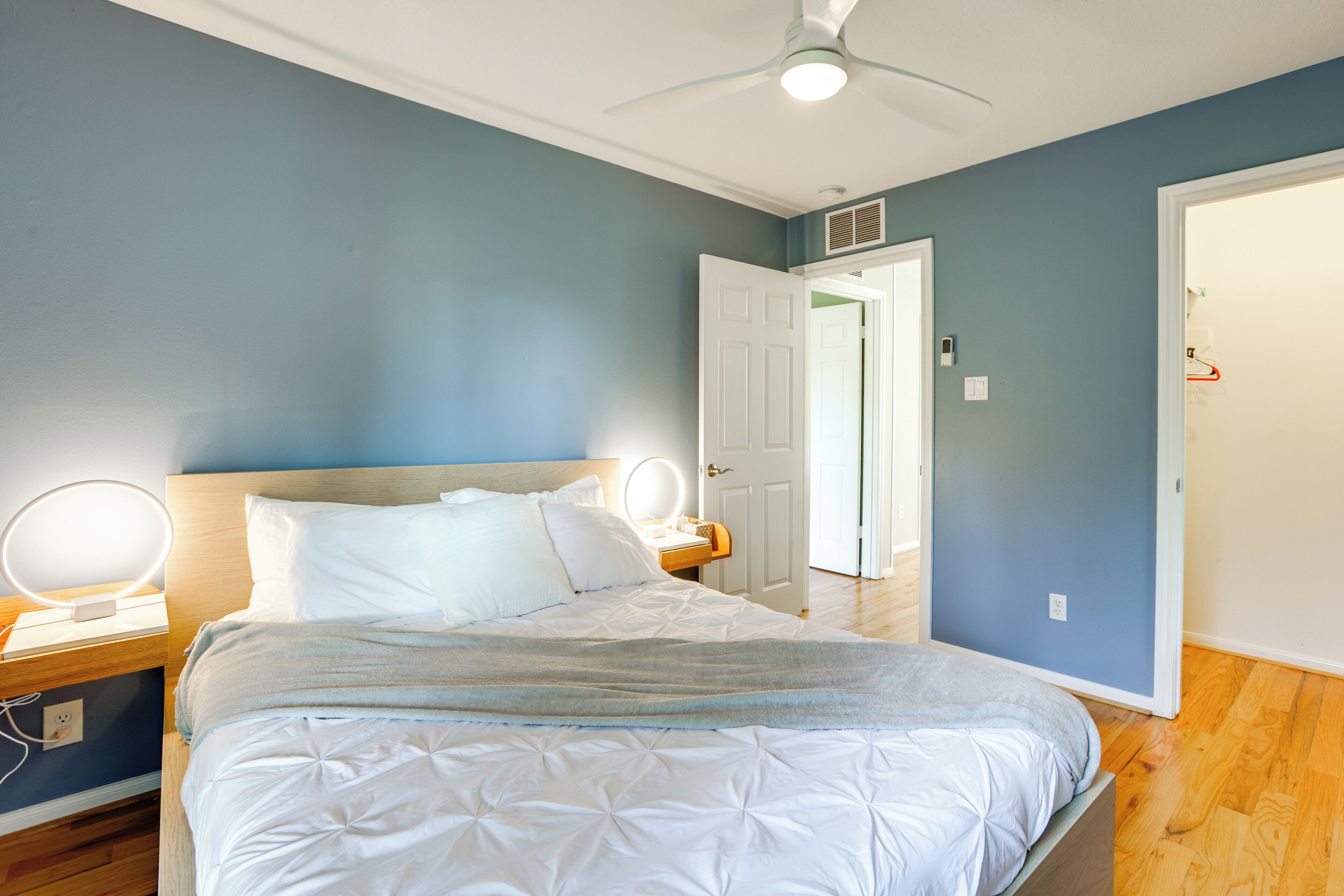 A bedroom with a bed, bedside lamps, a ceiling fan, and a door leading to another room.