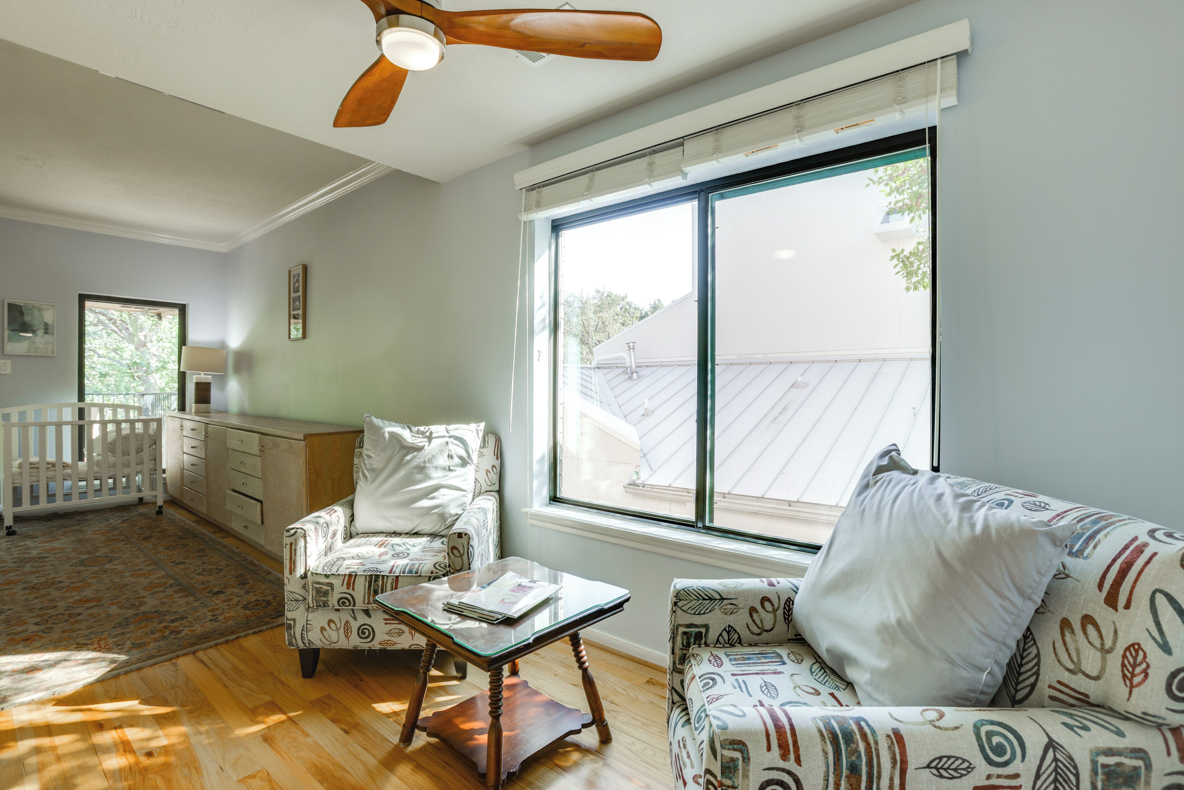 Un salon spacieux avec une grande fenêtre, un ventilateur de plafond, un berceau blanc et des meubles à motifs.