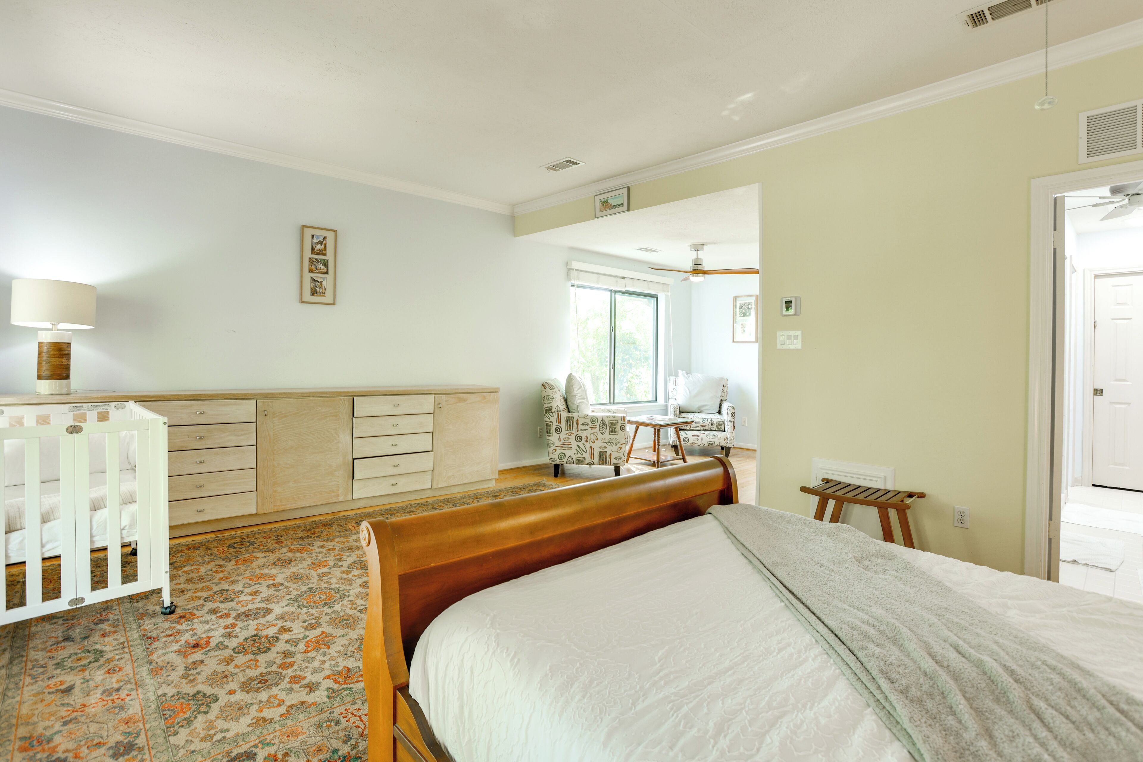 Une chambre à coucher avec un grand lit, une tête de lit en bois, un petit bureau, une chaise, un berceau et une fenêtre avec des stores.