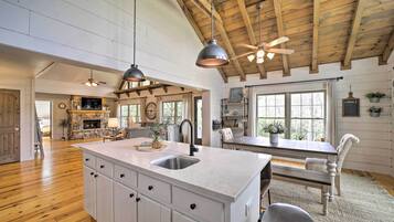 A spacious kitchen with a large island, wooden ceiling beams, pendant lighting, and a stone fireplace.