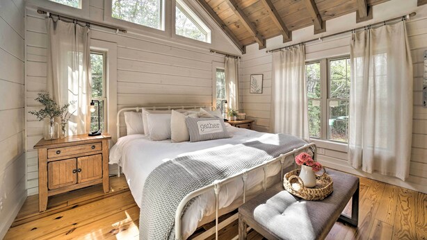 A bedroom with a wooden bed, a nightstand, a bench, and large windows. - Deck + Fire Pits: Lake Toxaway Getaway! (Lake Toxaway)