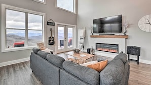 Ein geräumiges Wohnzimmer mit einer großen, grauen Eckcouch, einem Flachbildfernseher über einem Kamin und großen Fenstern mit Blick auf die Berge.