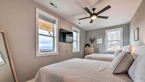 Ein Schlafzimmer mit zwei Betten, einem Deckenventilator, einem an der Wand befestigten Fernseher und großen Fenstern.
