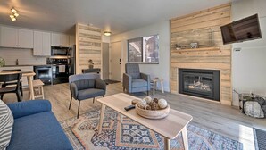 A modern living room with a fireplace, a TV mounted on the wall, and a kitchen area with white cabinets and a black refrigerator. - Renovated Park City Condo: Hike, Bike & Ski! (Park City)
