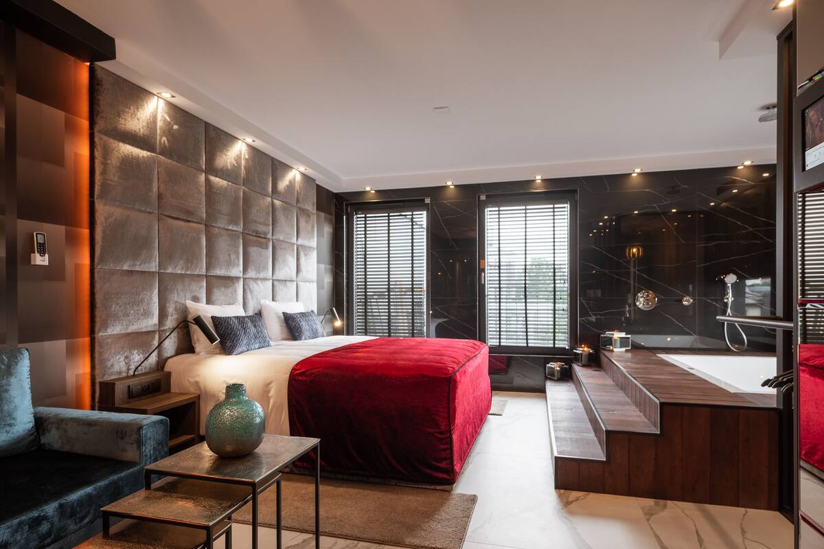 A modern bedroom with a large bed, a red accent wall, a built-in shower, and a small seating area.