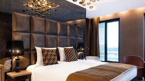 A modern hotel room with a large bed, a headboard with a geometric pattern, a bedside table with a lamp, and a view of the city through large windows. - Art Suites Luxury Hotel (Kraków)