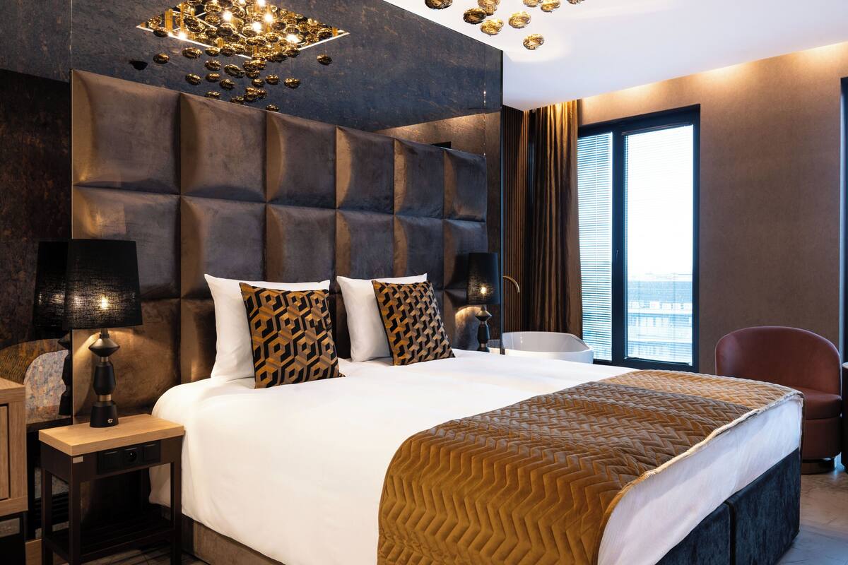 A modern hotel room with a large bed, a headboard with a geometric pattern, a bedside table with a lamp, and a view of the city through large windows.