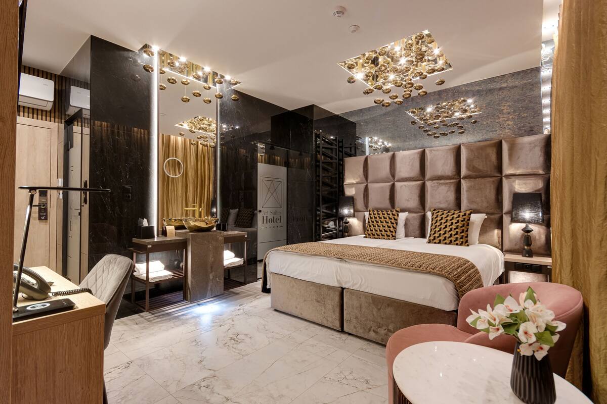 A modern hotel room with a large bed, a desk with a telephone, a chair, a vanity with a mirror, and a decorative wall panel.