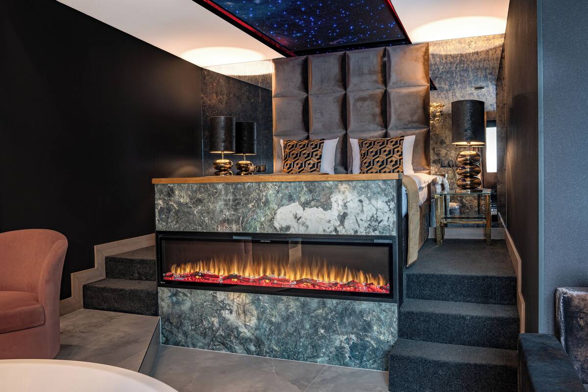 A modern bedroom with a fireplace, a bed with patterned pillows, and a night sky ceiling design.
