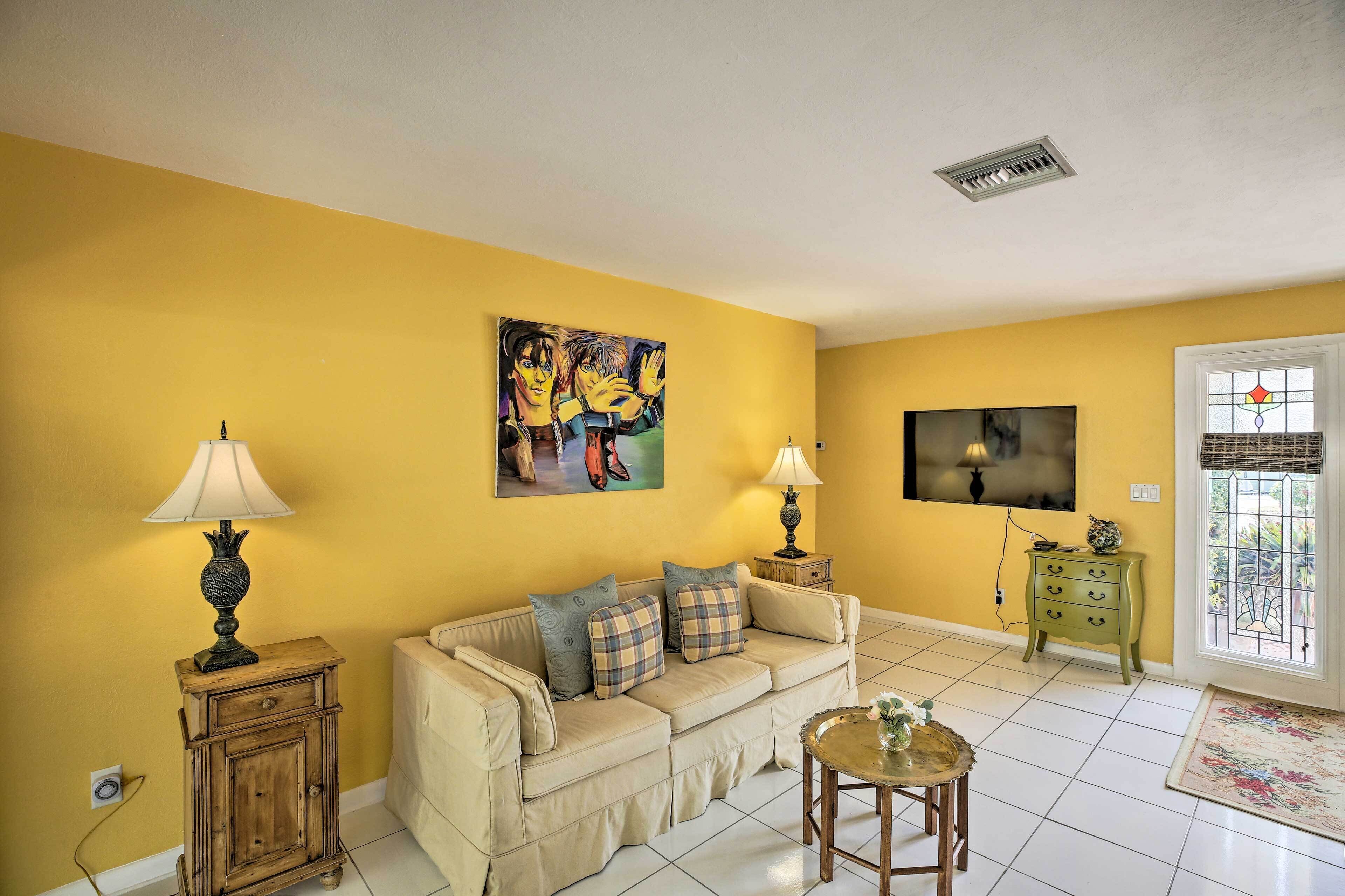 A living room with a yellow wall, a beige sofa, two side tables with lamps, a painting, a television, and a window with a view.