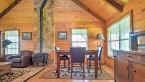 A cozy cabin interior with a stone chimney, wooden walls, a fireplace, a dining area with chairs, and a large window with blinds. - On Farm: Rustic Carmine Log Cabin w/ Porch! (Carmine)