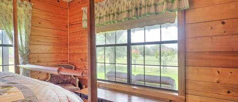 A wooden cabin room with a bed, a bench, and a window with a view of a green landscape.