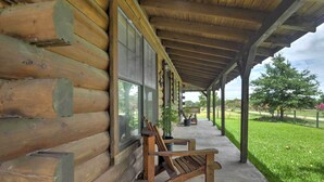 A log cabin with a covered porch, wooden benches, and a view of a well-maintained lawn and trees. - On Farm: Rustic Carmine Log Cabin w/ Porch! (Carmine)