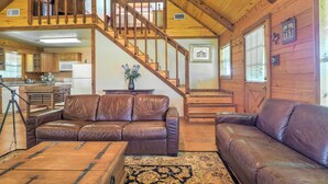A spacious living area with a wooden staircase, leather sofas, and a kitchen in the background. - On Farm: Rustic Carmine Log Cabin w/ Porch! (Carmine)