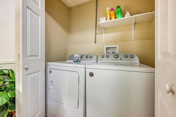 A laundry area with two washers and dryers, a shelf with cleaning supplies, and a door with a plant visible.