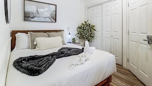 Una cama bien hecha con sábanas blancas y un plaid negro, una lámpara de noche y una planta en maceta sobre una mesita de noche.