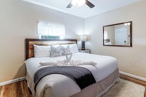 Une chambre d’hôtel avec un lit, une table de chevet, une lampe, un ventilateur de plafond, une fenêtre avec des rideaux et un miroir au mur.