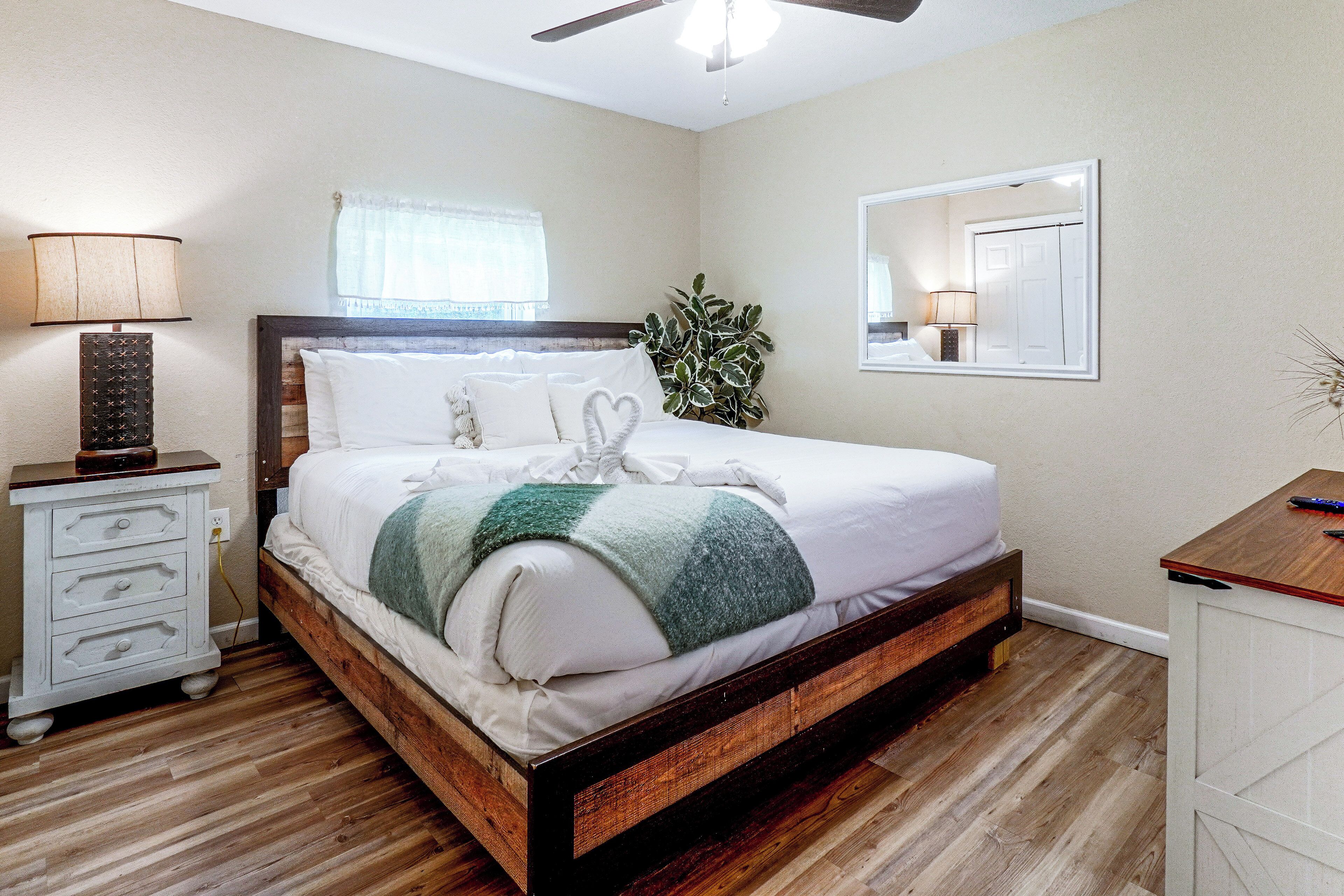 A bedroom with a wooden bed frame, white bedding, a bedside lamp, a mirror, and a plant.