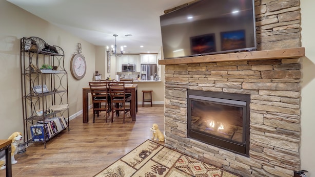 A living room with a stone fireplace, a flat-screen TV mounted above it, a dining area with a wooden table and chairs, and a bookshelf with decorative items. - Mtn-view Condo w/ Lookout Deck in Banner Elk! (Banner Elk)