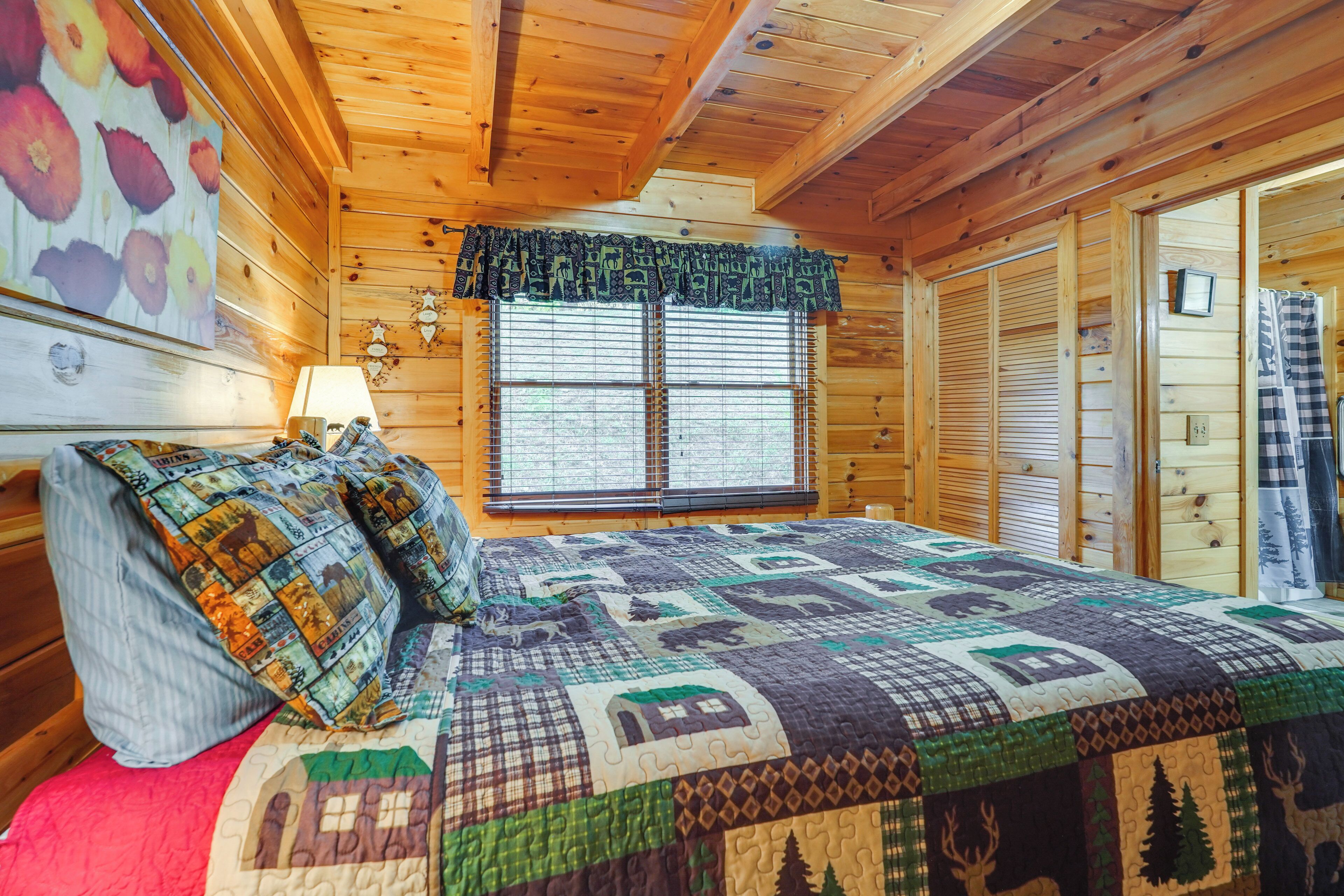 A wooden cabin bedroom with a quilted bedspread, a window with blinds, and a wall-mounted lamp.