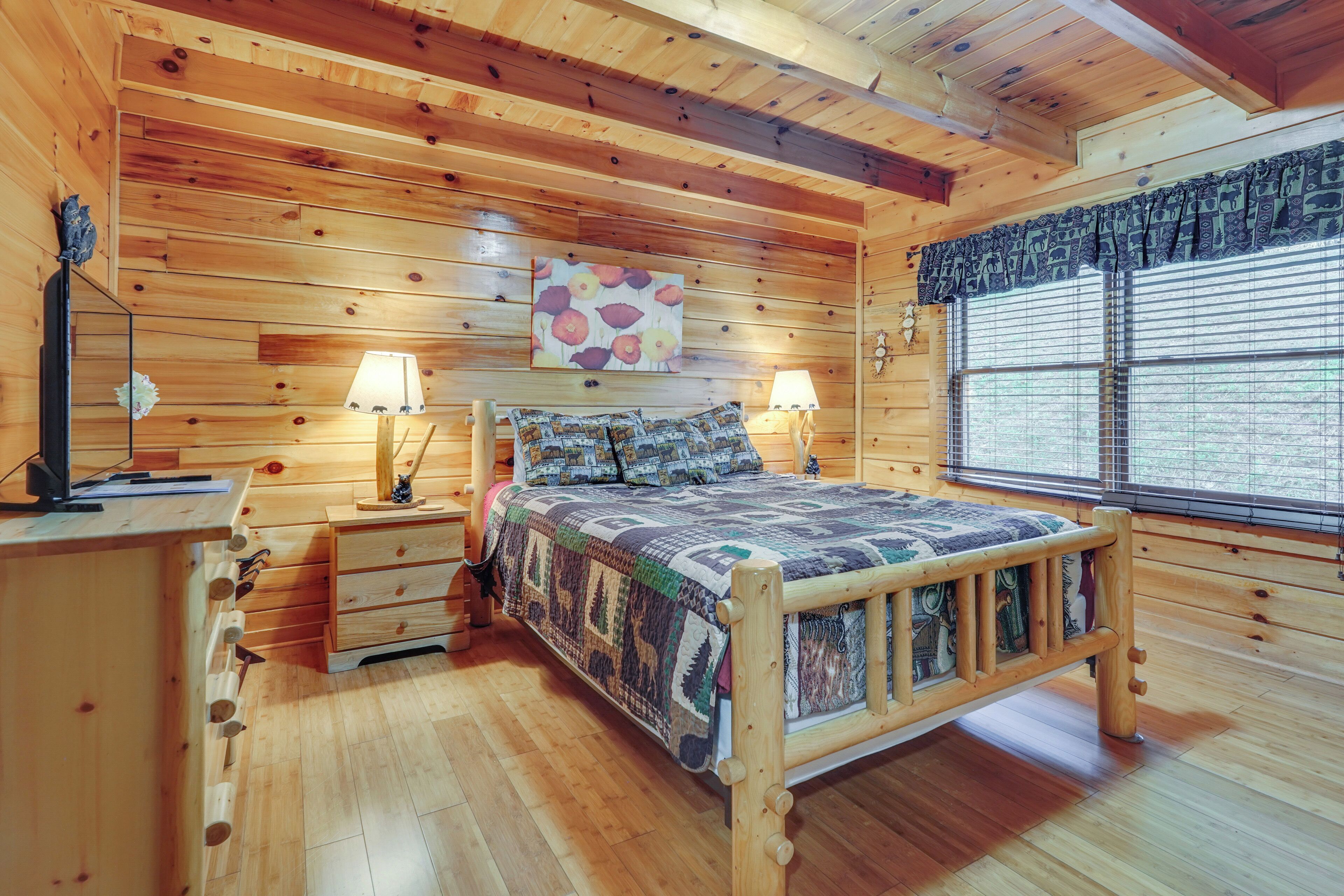 Une chambre à coucher dans un chalet en bois, avec un lit, deux tables de chevet, une petite table, une fenêtre avec des stores et un tableau accroché au mur.