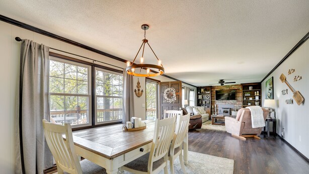 A dining area with a wooden table, white chairs, and a chandelier. There is a large window with curtains, a fireplace, and a television in the background. - Charming Shell Knob Home: 16 Mi to Dogwood Canyon! (Shell Knob)