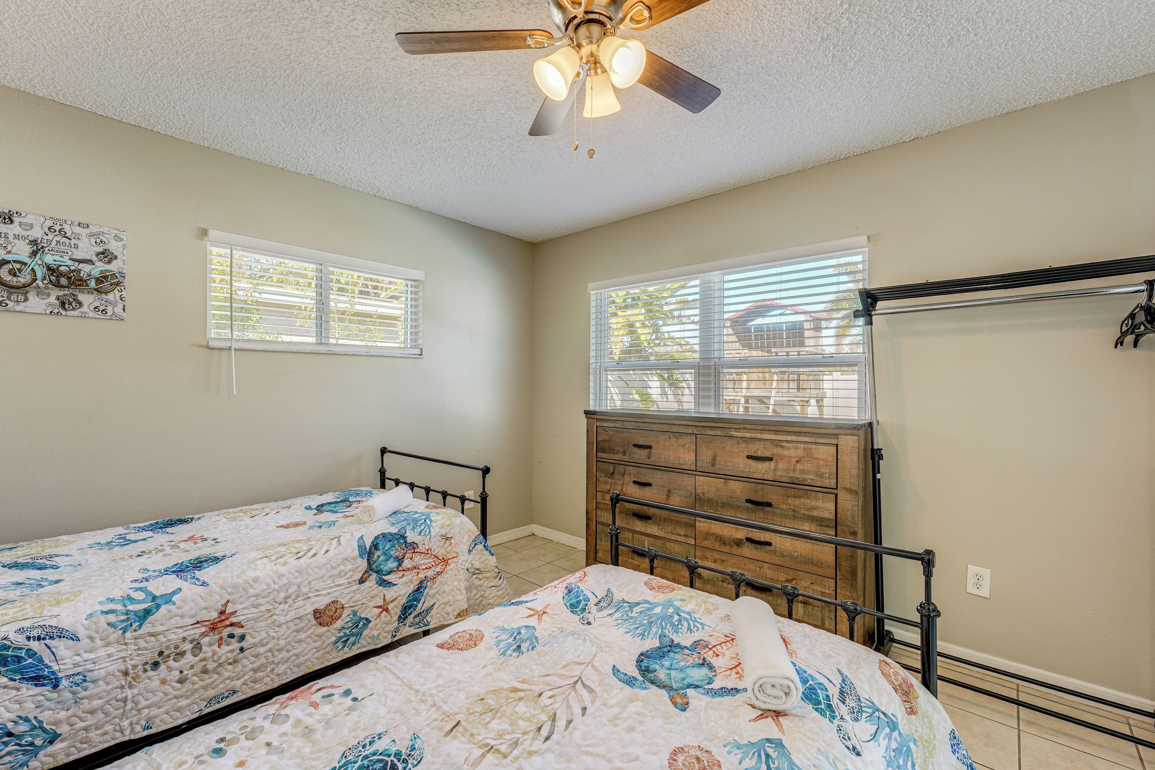 A bedroom with two beds, a ceiling fan, a dresser, and two windows with blinds.