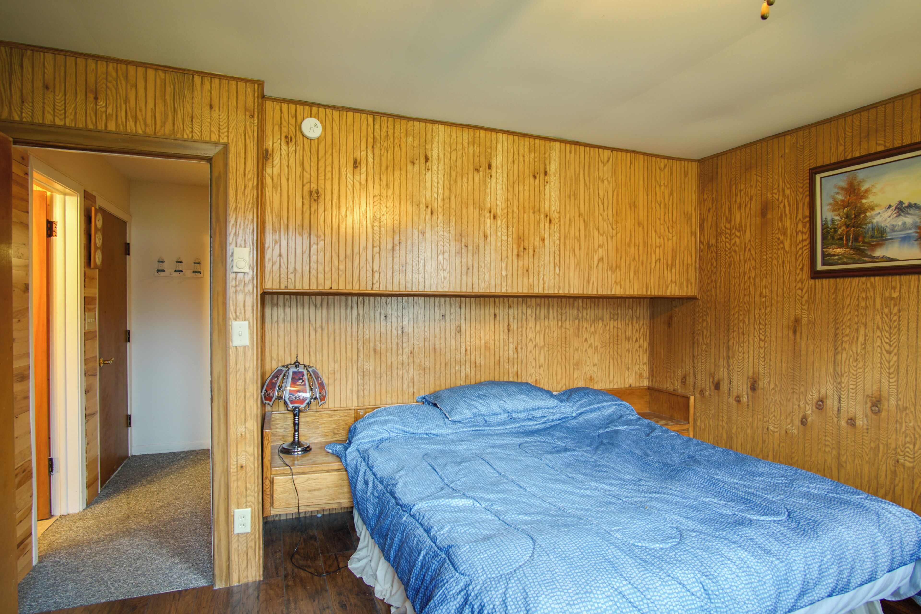 A bedroom with wooden paneling, a bed with a blue quilt, a nightstand with a lamp, and a painting on the wall.