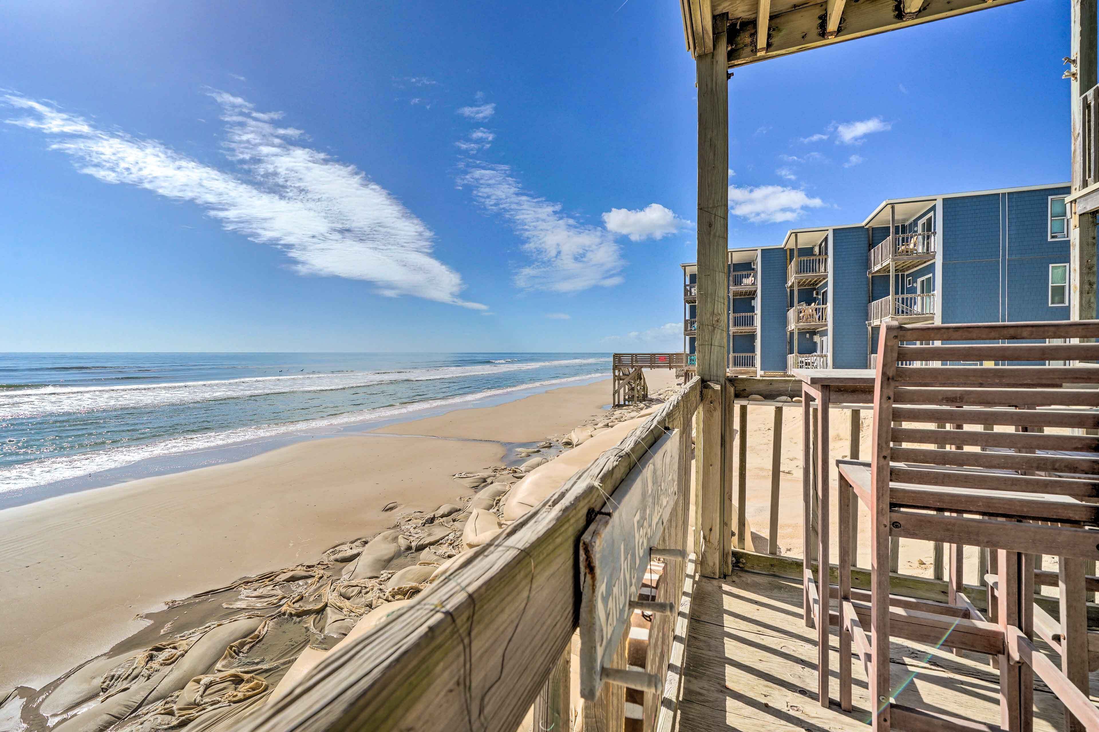 A beach view from a balcony with a wooden railing, overlooking a sandy beach and the ocean.