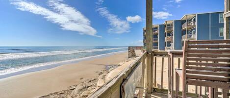 A beach view from a balcony with a wooden railing, overlooking a sandy beach and the ocean.