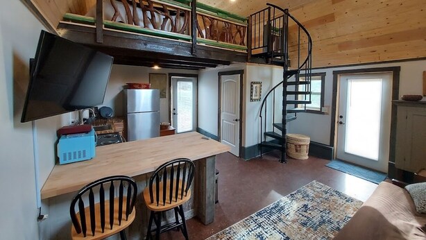 A cozy cabin interior with a wooden ceiling, a dining area with two chairs, a kitchen with a stove and refrigerator, a staircase, and a television mounted on the wall. - Rustic Hendersonville Farm Studio w/ Private Deck (Hendersonville)