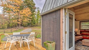 A wooden deck with a table and chairs, a potted plant, and a sliding glass door leading to a room with a red chair and a lamp. - Rustic Hendersonville Farm Studio w/ Private Deck (Hendersonville)