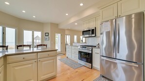 Una cocina amplia con gabinetes blancos, electrodomésticos de acero inoxidable y ventanas grandes que ofrecen vistas al exterior.