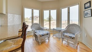 Una habitación amplia con cocina, dos sillones de mimbre con cojines azules, una mesita y grandes ventanales con vistas a la playa.