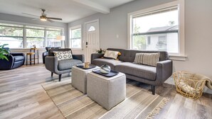 A modern living room with a grey sofa, two ottomans, a coffee table, and a wicker basket. There are large windows, a ceiling fan, and a door with a stained glass window. - Dtwn Winter Garden Home: 12 Mi to Disney! (Winter Garden)