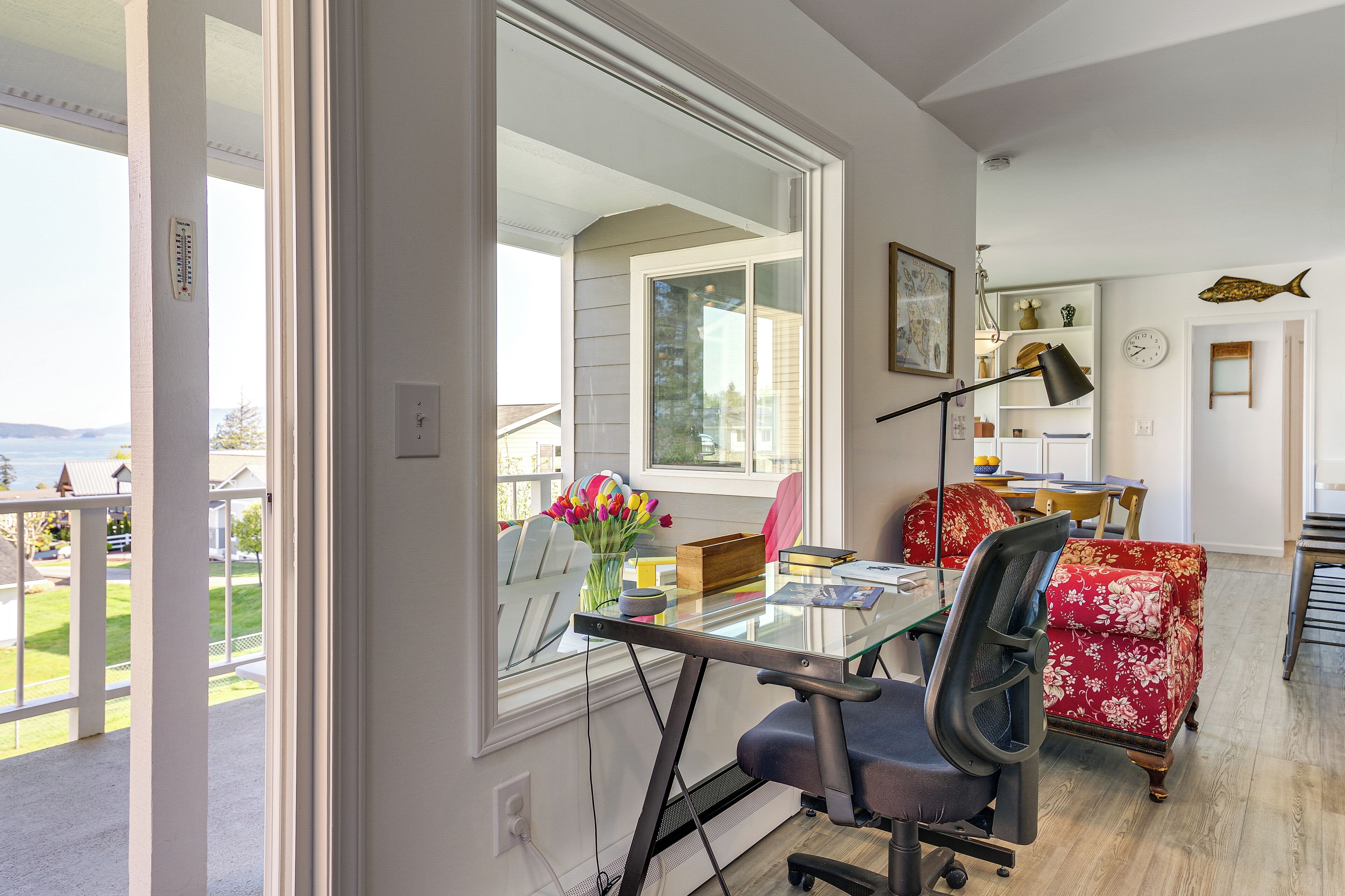 A modern home office with a glass desk, a red floral armchair, and a view of the outdoors.