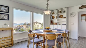 Un coin repas avec une table ronde en bois, six chaises et une vue sur les maisons à travers de grandes fenêtres.