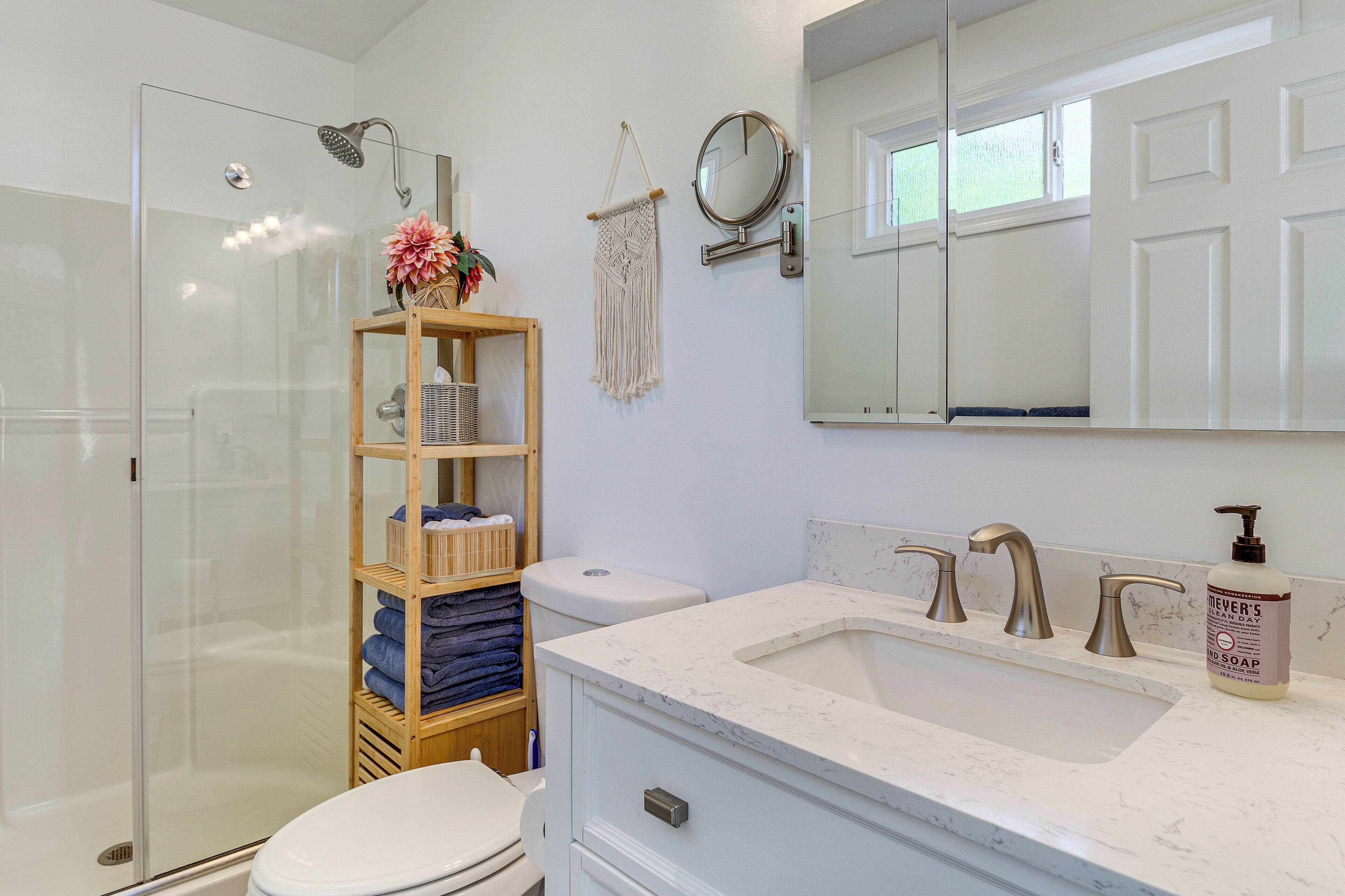 A modern bathroom with a glass shower enclosure, a wooden shelf with towels, a white sink with a marble countertop, and a round mirror above the sink.