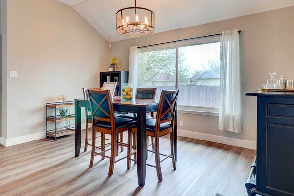 Un comedor con mesa y sillas, una araña, una ventana con cortinas y vistas al exterior.