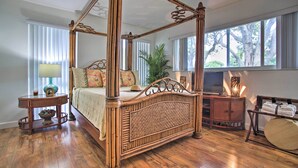 Un lit à baldaquin avec une tête de lit en osier, une table de chevet avec une lampe, et une vue sur l’extérieur grâce à de grandes fenêtres.