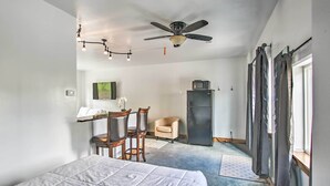 Een slaapkamer met een bed, een plafondventilator, een bureau met stoel, een fauteuil, een koelkast en een raam met gordijnen.