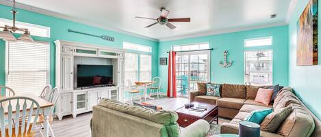 Un salon aux murs de couleur turquoise, doté d’un ventilateur de plafond, d’un téléviseur, d’un canapé et d’un coin repas avec des chaises blanches.