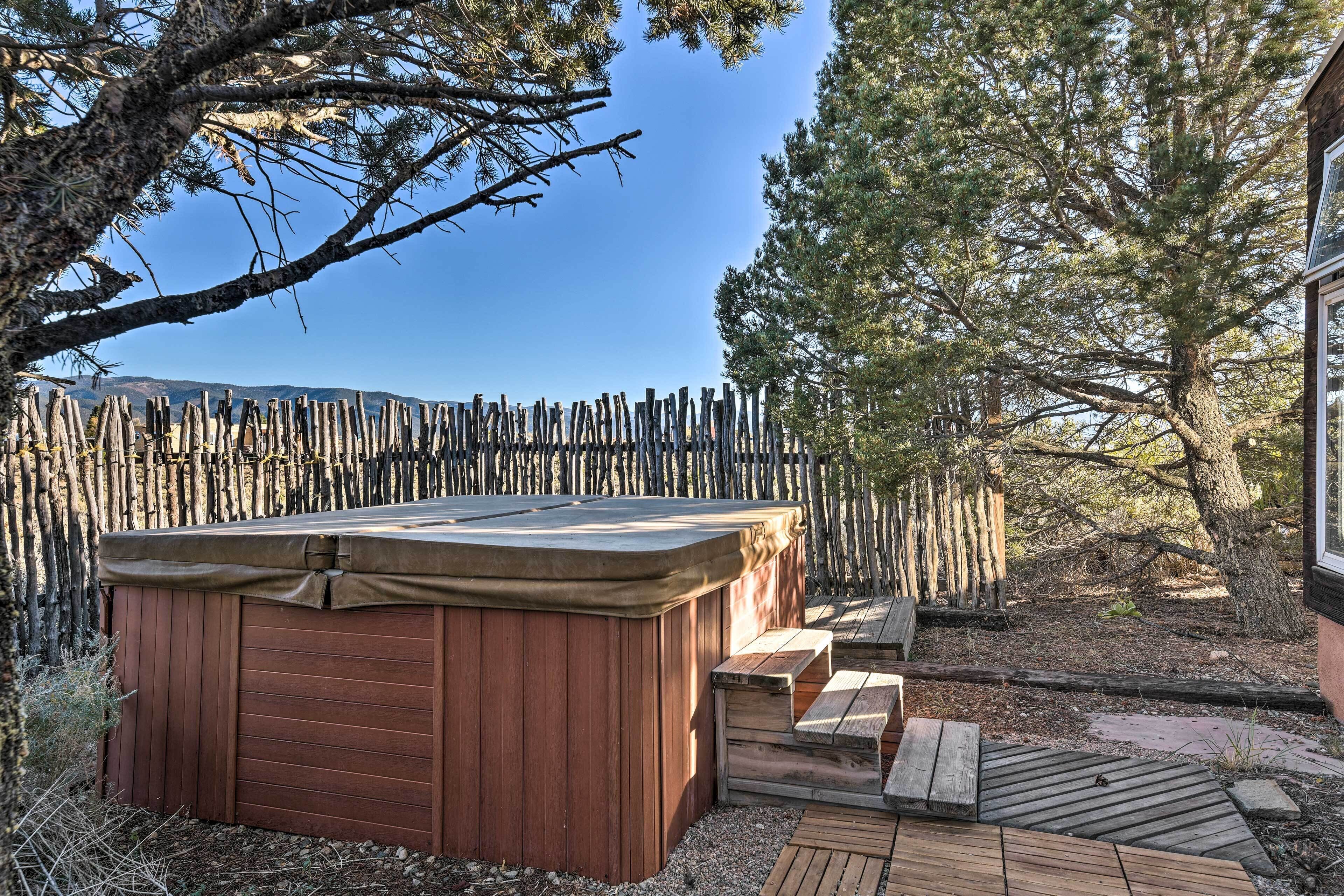 A wooden hot tub with a bench and a wooden deck in a backyard with a wooden fence and trees.