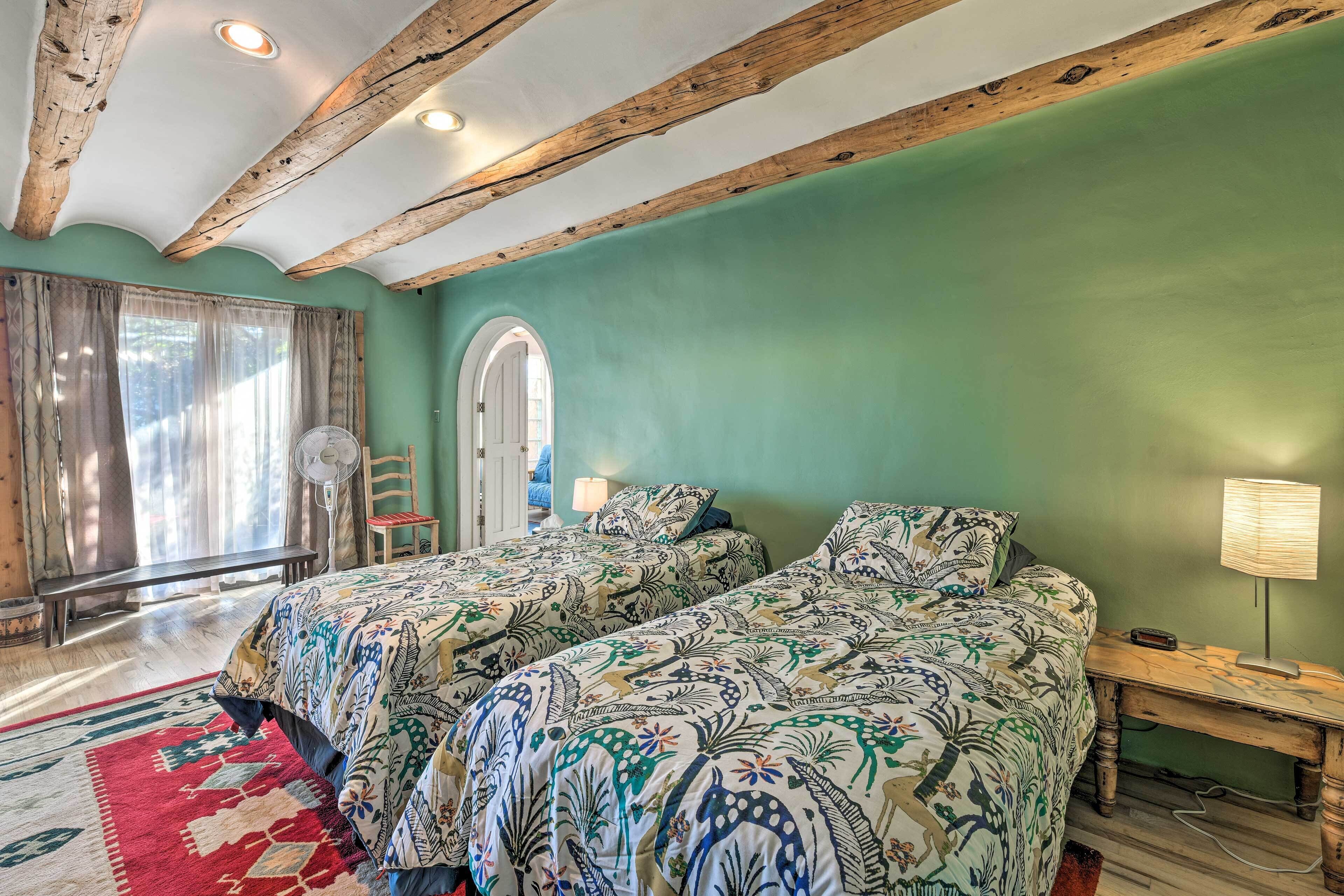A bedroom with two beds, a wooden ceiling, a green wall, a window with curtains, a fan, and a nightstand with a lamp.