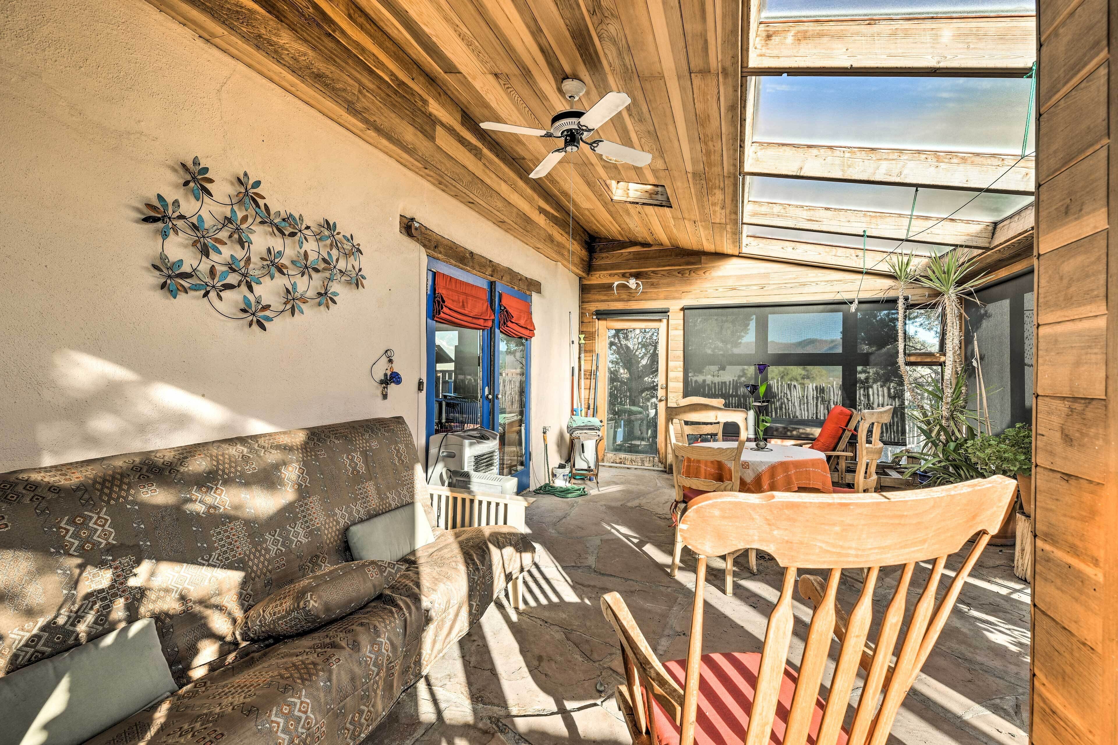A sunlit patio with a wooden ceiling, a ceiling fan, a wall-mounted decorative metal piece, a wooden chair, and a table with a red cushion.