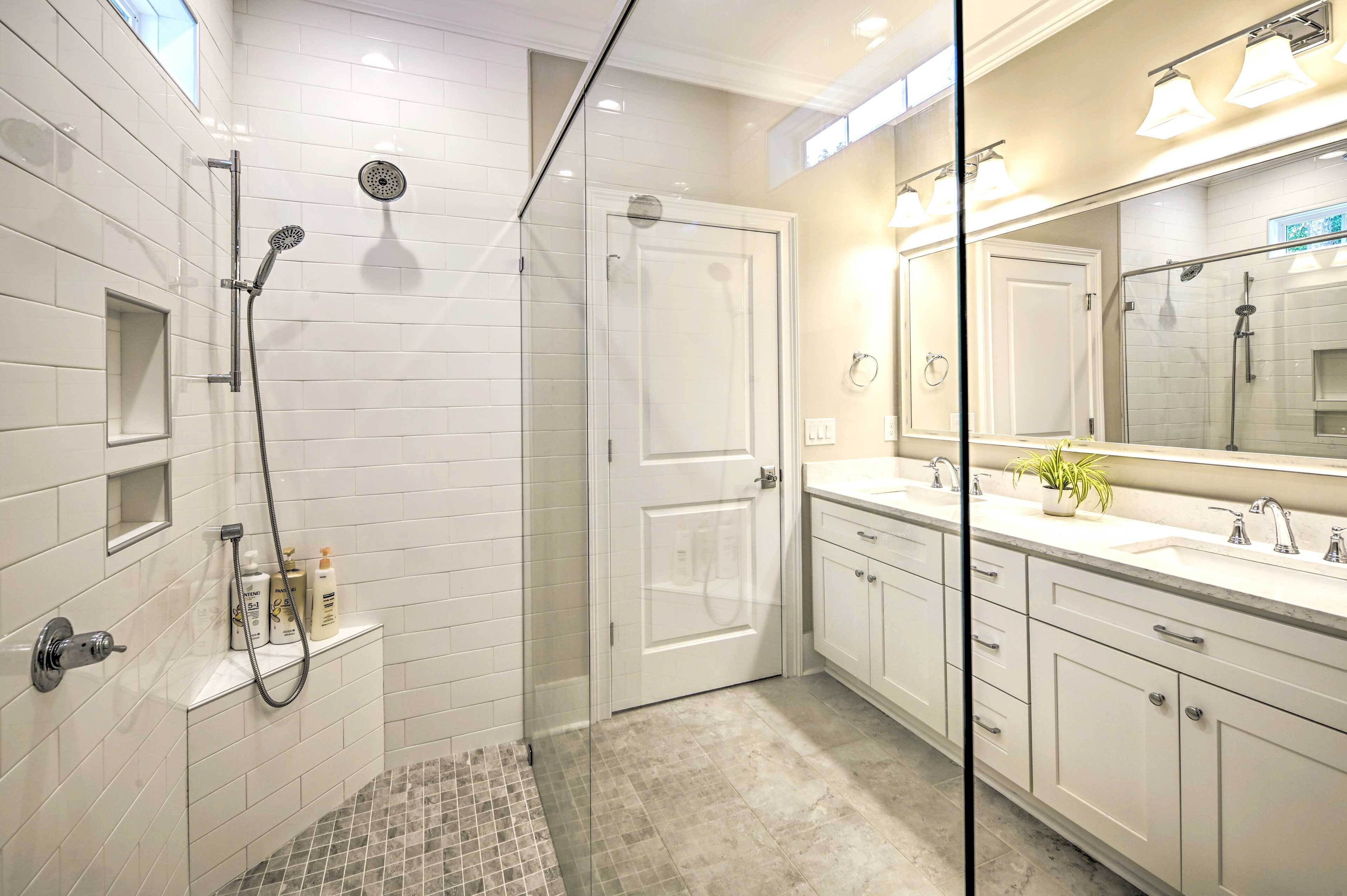 Un baño moderno con ducha envuelta en vidrio, paredes cubiertas de azulejos blancos, un espejo grande y un lavamanos con lavabo y encimera.